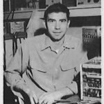 Black and white photo of a man sitting indoors near electronic equipment.